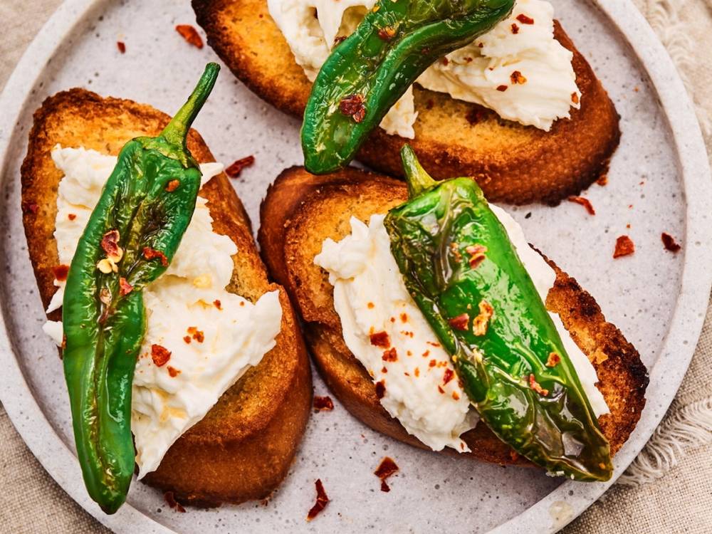 Tre toast med kremost og grillede grønne chili på en tallerken, pyntet med chili flakes.