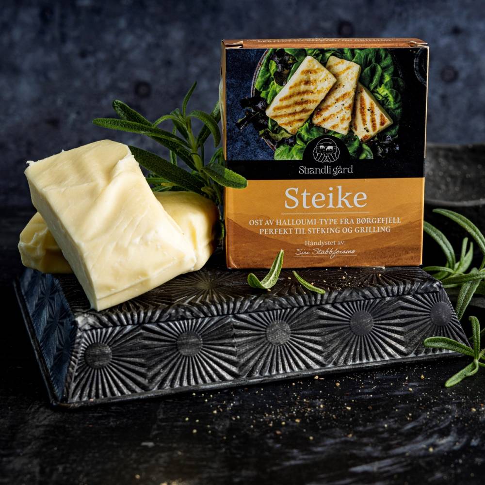Pakke med Steike ost fra Strandli Gård, plassert ved siden av et svart ostebrett og noen grønne urter på en mørk overflate.