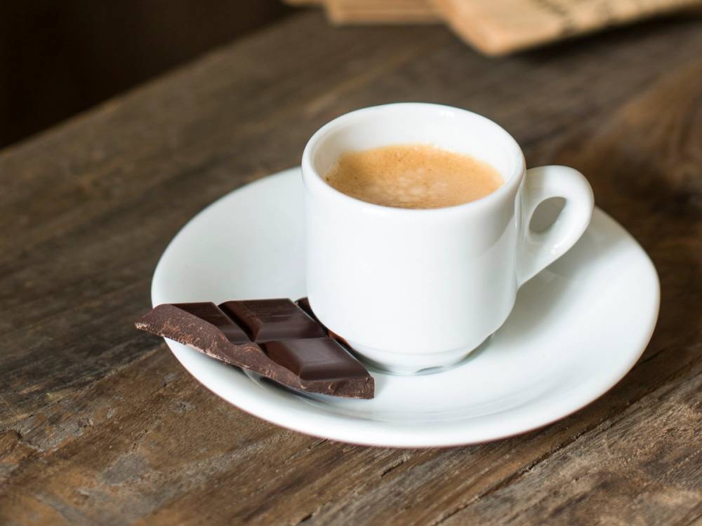 En kopp espresso på en hvit tallerken med et stykke sjokolade ved siden av, plassert på et trebord.
