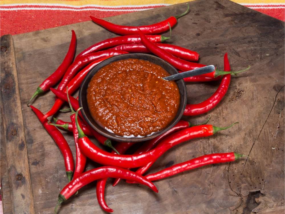 En skål med chilimani saus omgitt av mange friske røde chilipepper på en treplate
