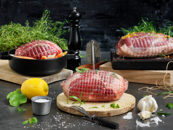 Ulike rå kjøttstykker på skjærebrett og panner med urter og krydder, klar for tilberedning, på et kjøkkenbord.