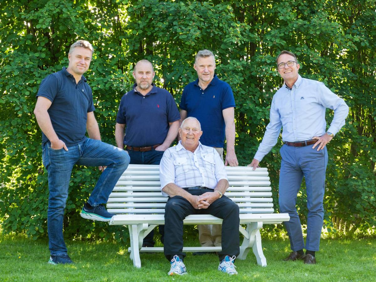 Fem menn med bløte klær og jeans står rundt en hvit benk i en grønn park, hvor en mann sitter på benken.