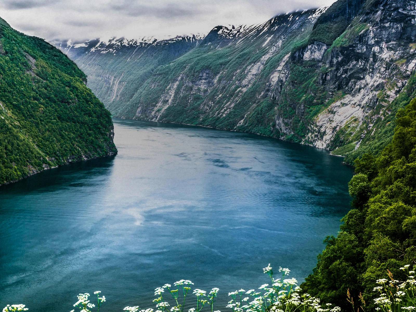 En innsjø omgitt av høye, grønne fjell med skyer over fjelltoppene