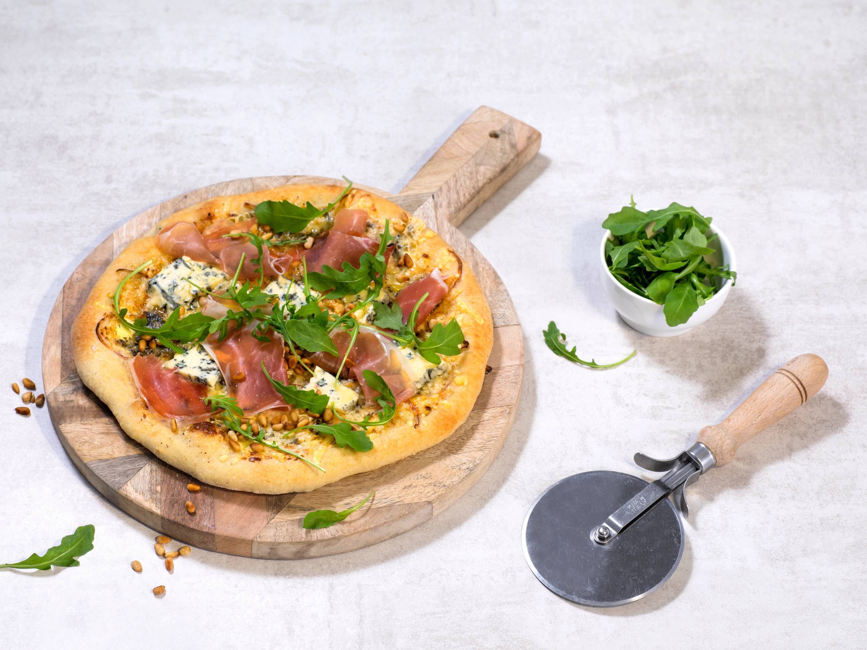 Pizza med blåmuggost, skinke og ruccola på en treplate med en liten bolle med ruccola ved siden av.