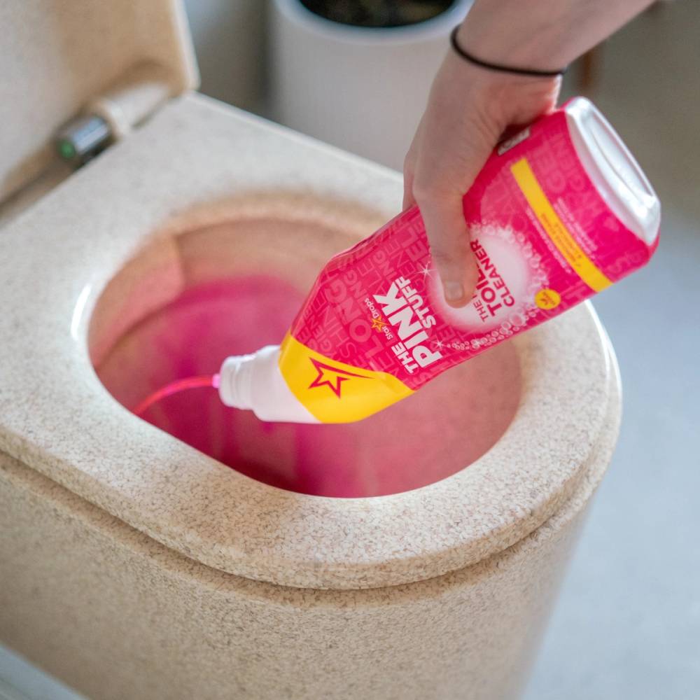 En person bruker en rosa rengjøringskrem fra The Pink Stuff til å rengjøre en overflate, med en hånd som holder tuben over et hull i en keramisk overflate.