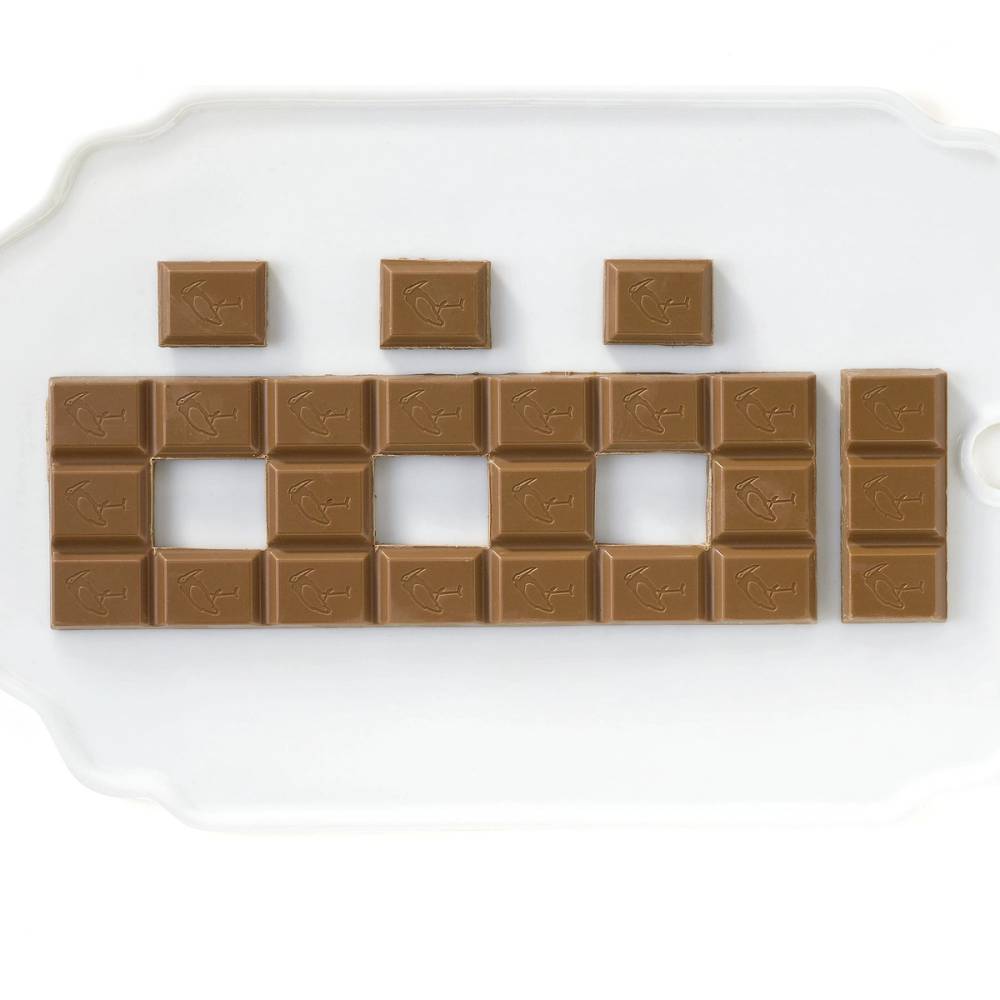 En rekke med kvadratiske sjokoladebiter og noen større sjokoladestykker plassert på et hvitt underlag, hvor noen av sjokoladene er stablet for å danne et huslignende byggverk.