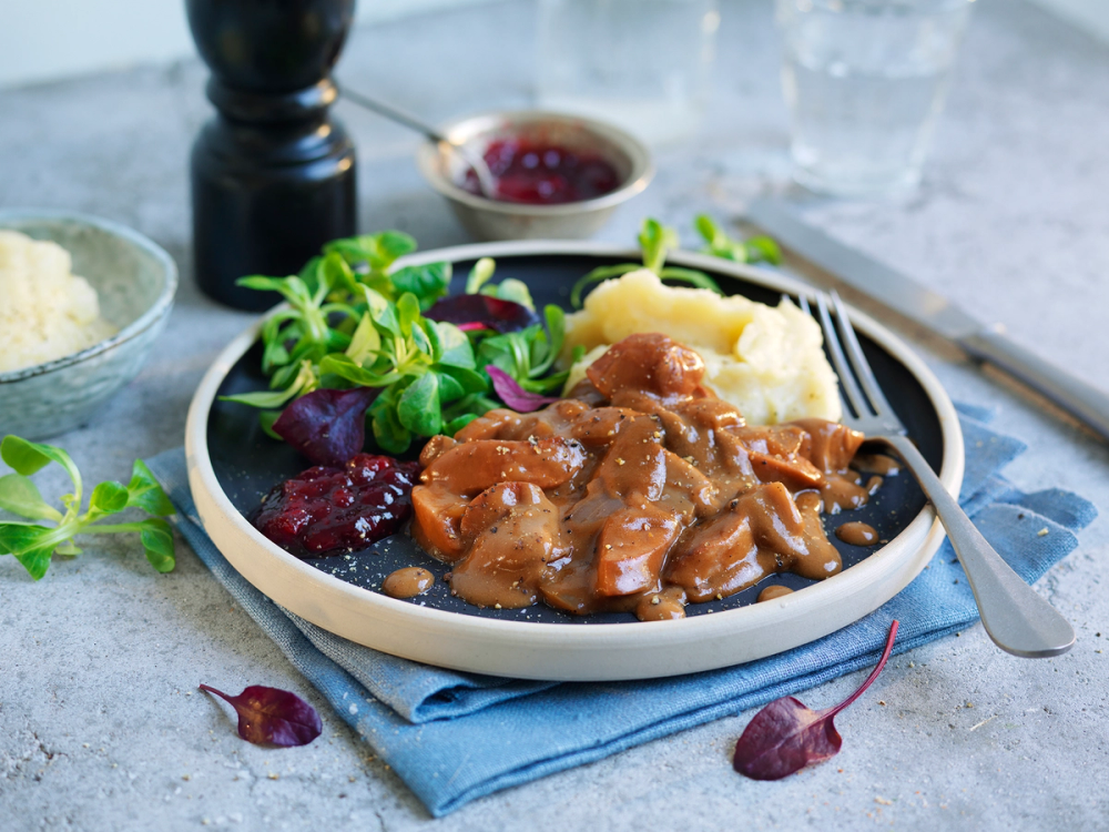 Stroganoff med pølsebiter servert med potetmos og en liten skål med tyttebærsyltetøy på et bord.