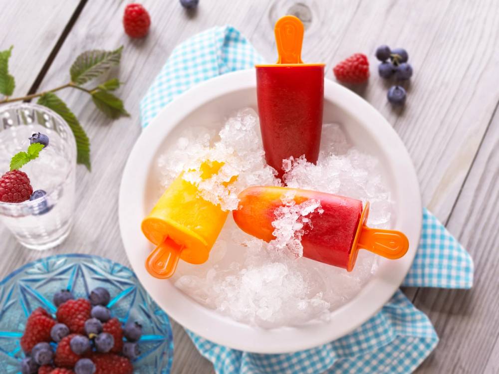 Hjemmelaget iskrem i tre ulike farger på isformer, plassert i en bolle med knust is, omgitt av bær og et glass med bær ved siden av.