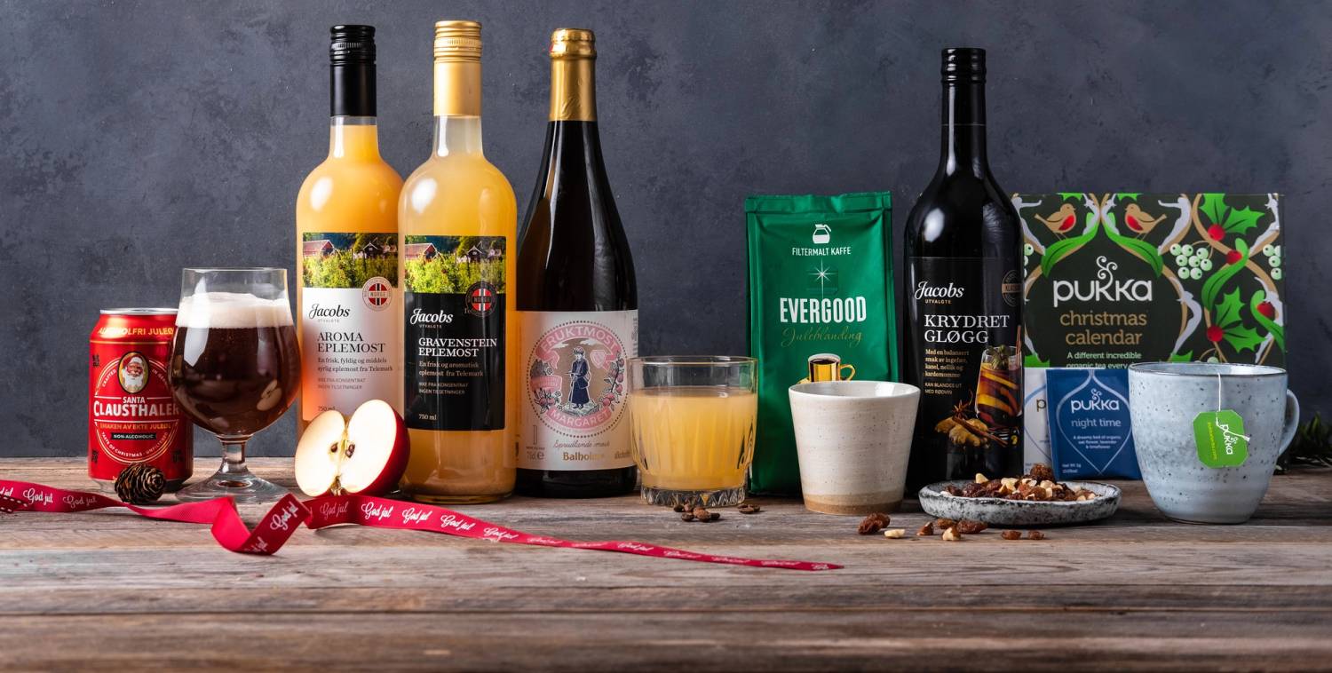 Et utvalg av juledrikker, inkludert vin, øl, brusbokser og te, på et trebord med julekule og pepperkaker i bakgrunnen.
