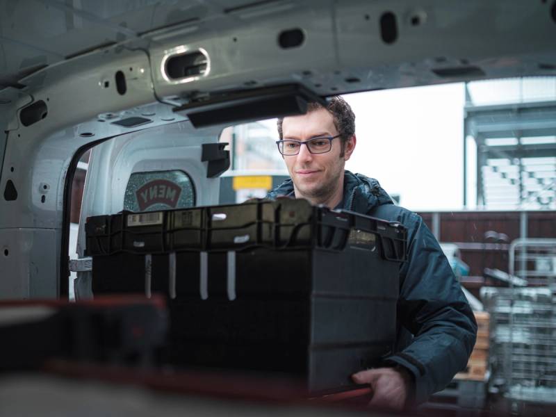 En person med ansiktet dekket av en pikselert maske som står inne i en flykropp og håndterer en stor svart koffert, med flyets interiør rundt seg.