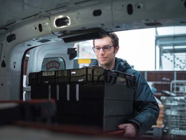 En person med ansiktet dekket av en pikselert maske som står inne i en flykropp og håndterer en stor svart koffert, med flyets interiør rundt seg.