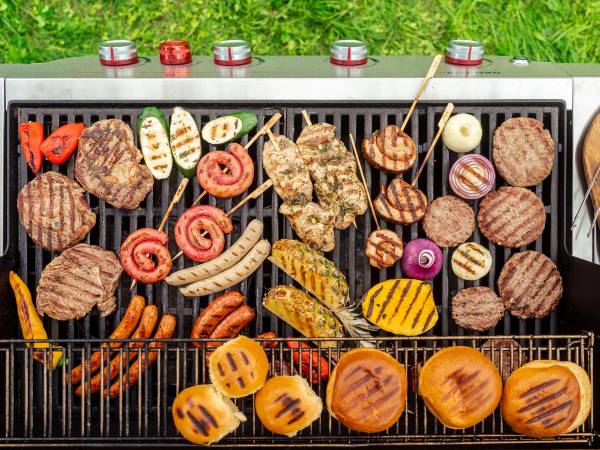 Grill med ulike kjøtt- og pølsevarer, grønnsaker og grillspyd på rist, med gress og blomster i bakgrunnen.