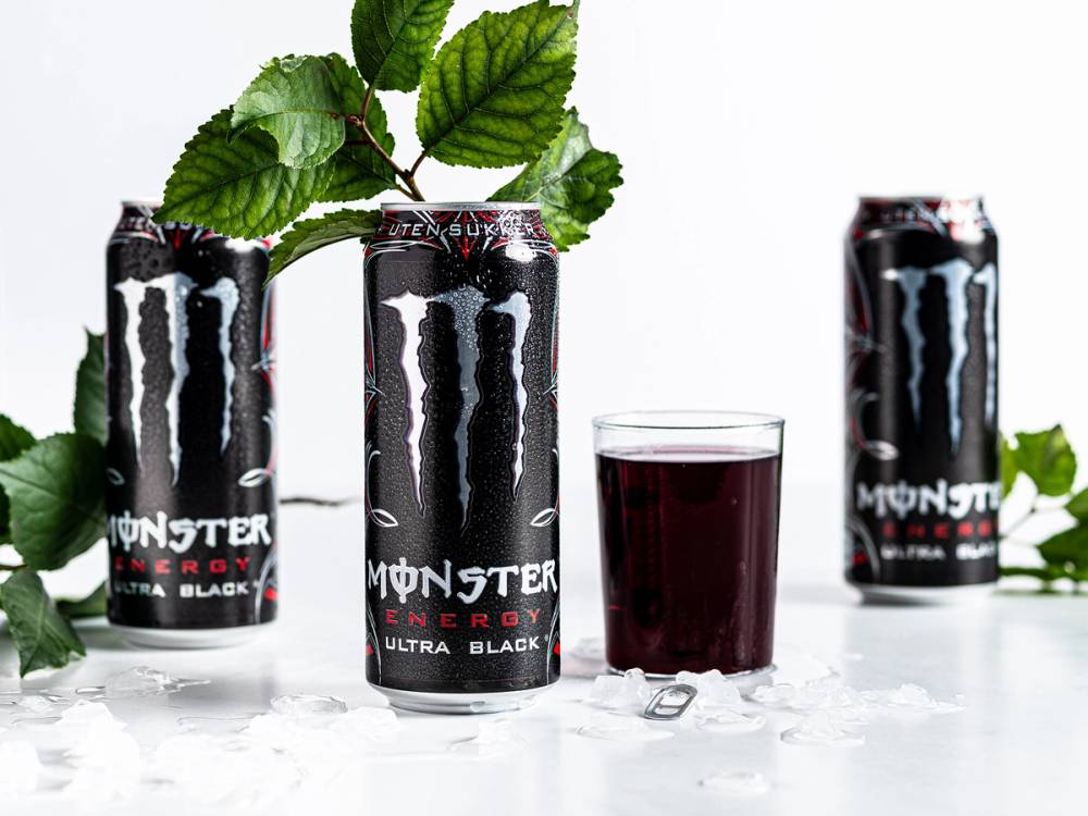 Tre Monster Energy Ultra Black energidrikker og et glass med mørk drikke, med grønne blader i bakgrunnen.