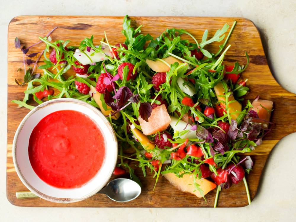 En frisk salat med melon, bringebær, ruccula og bringebærdressing på en treplate.