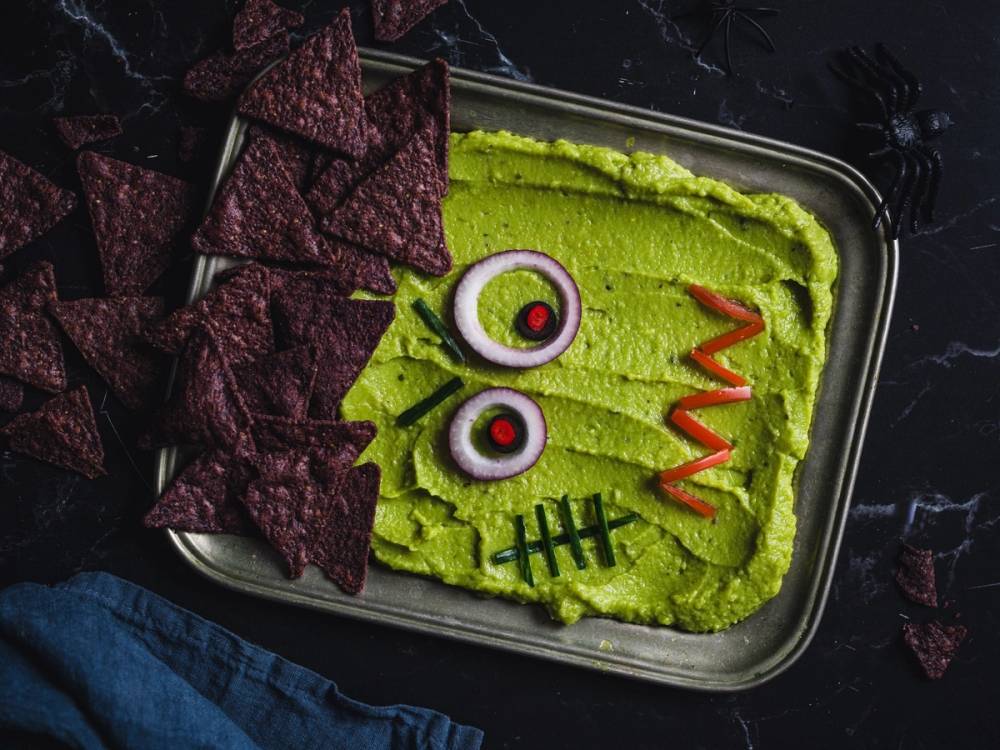 En skål med grønn hummus dekorert med ansikt, øyne, munn og sår, omgitt av tortillachips, på en svart bakgrunn.