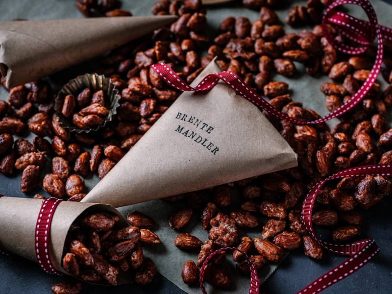 Brente mandler i en papirpose med en rød og hvit stripet sløyfe, omgitt av flere mandler på en mørk bakgrunn.
