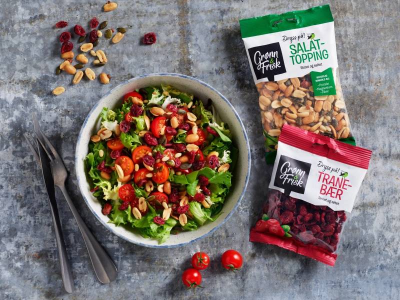 En salat med ulike grønnsaker og nøtter i en hvit bolle, med to poser med salat-topping og tranebær ved siden av på en grå overflate.