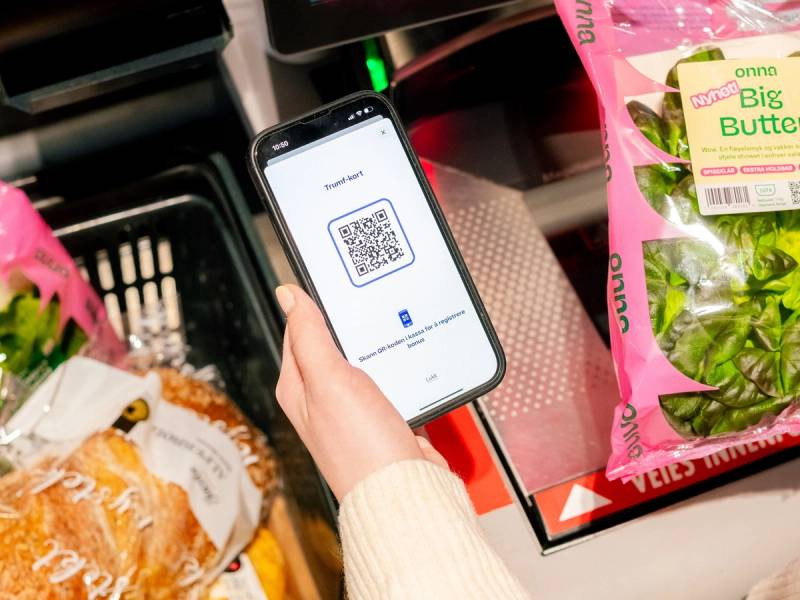 En person skanner en QR-kode med en smarttelefon i en dagligvarebutikk, omgitt av grønnsaker og brød.