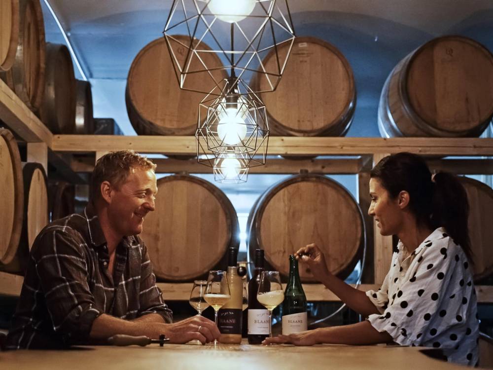 To personer sitter og snakker over et bord med vinflasker i en vinkjeller med store vinfat på veggen i bakgrunnen.