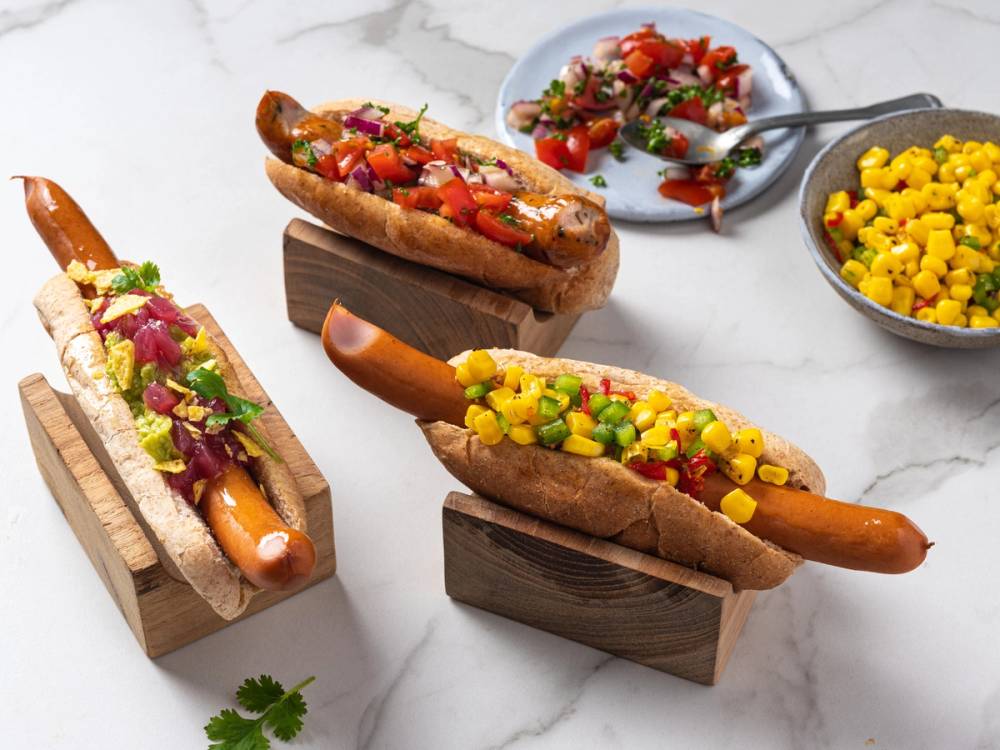 Tre pølser med sunn topping som er pyntet med grønnsaker, plassert på små trebrikker og servert med to små skåler med ekstra topping og en skje, på en hvit bakgrunn.