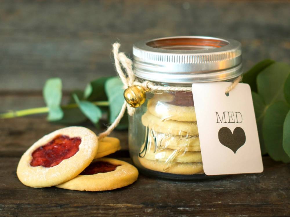 Småkaker med jordbærsyltetøy ved siden av et glass med kjeks og en tag med hjerte, på en treoverflate.