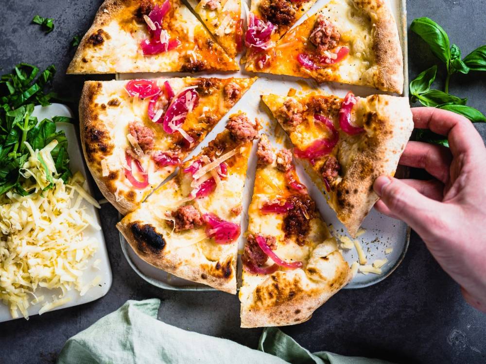 En person skjærer i en pizza med skinke og ost, plassert på et bord med en liten salat ved siden av.