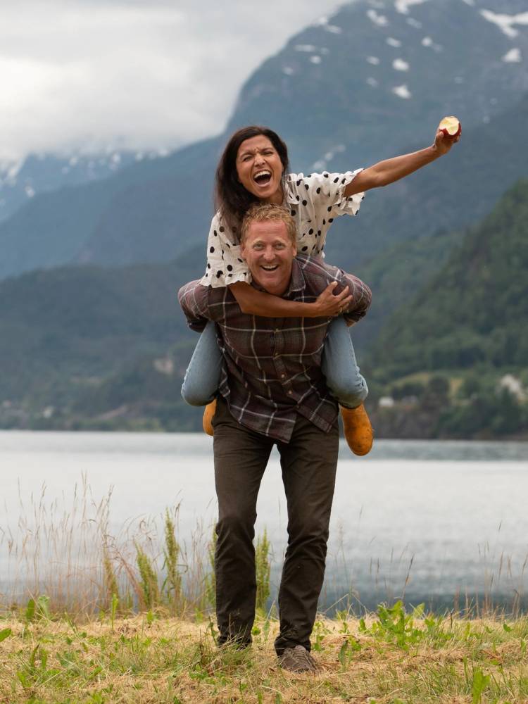 En voksen bærer en liten jente på skuldrene ved en innsjø med fjell i bakgrunnen, og jenta rekker ut armen med en liten gjenstand i hånden.