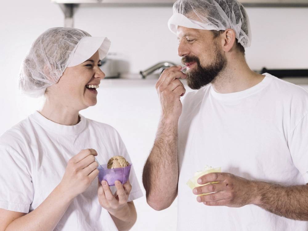 To personer i hvite klær og hårnett som smaker på mat i et kjøkkenmiljø, uten å vise ansiktene.