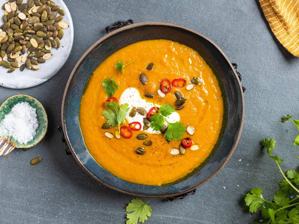 Gulrotsuppe servert i en svart bolle med pynt av chili, koriander og kokosflak, omgitt av ulike krydder og friske urter.