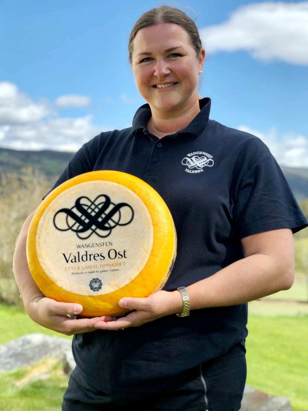 En person holder en stor ost med etiketten 'Valdres Ost' utendørs med grønne åser og blå himmel i bakgrunnen.