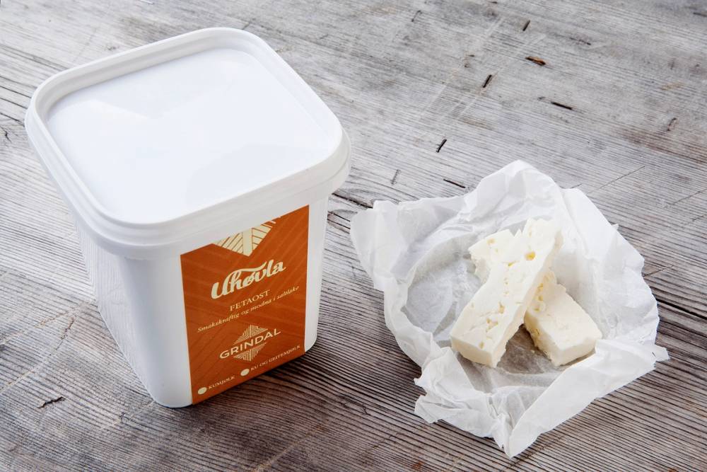 En bøtte med hvit ost og et stykke ost på et papir på en treoverflate.