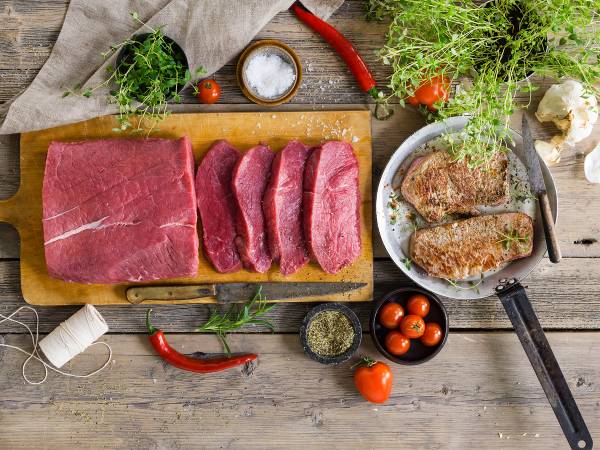Ulike typer av rått kjøtt på skjærebrett med urter, tomater og krydder rundt, klar for å bli stekt.