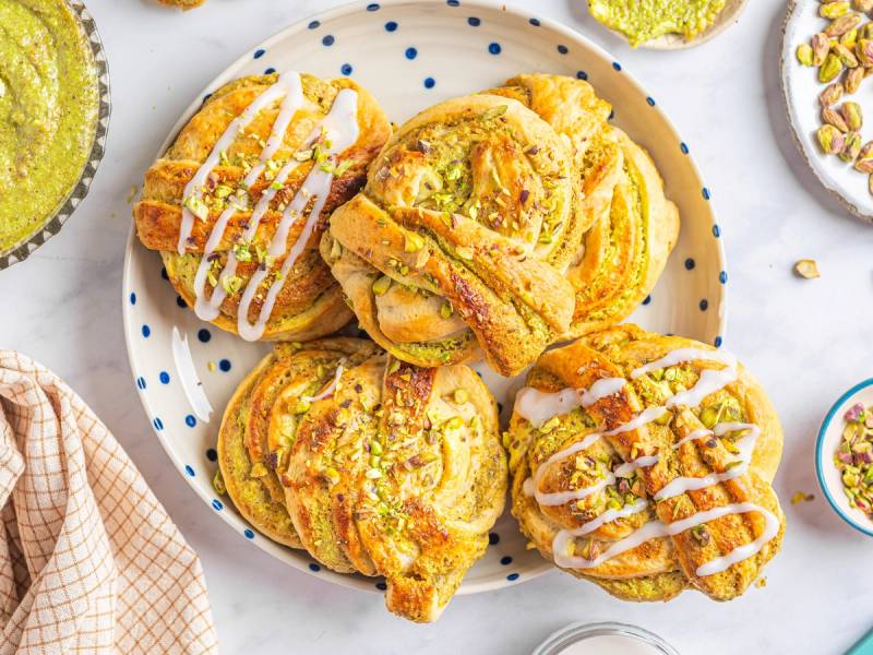 Pistasj-boller med hvit glasur på en hvit tallerken med blå prikker, omgitt av andre bakverk og små skåler med pynt.