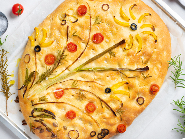 Focaccia med blomsterformede grønnsaker og cherrytomater pyntet på toppen, dekorert med urter rundt på en hvit overflate.
