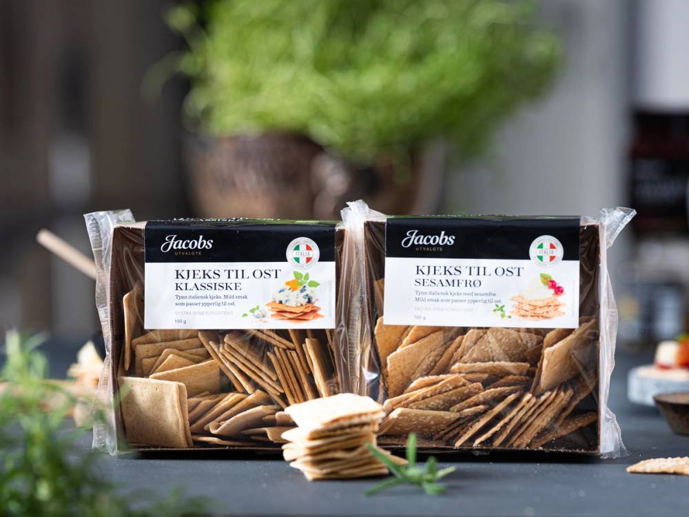 To poser med kjeks fra JU-kjeks-ost-10-nett, en med ostesmak og en med sesam, plassert på et bord med grønne planter i bakgrunnen.