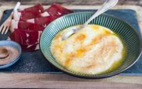 Rommigrot servert i en grønn bolle med en skje oppi, omgitt av skinke og salt på et trebord