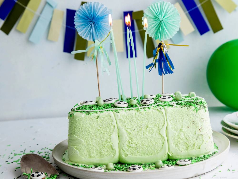 En grønn iskake dekorert med små fotballklistremerker og to telys på toppen, med bursdagsdekorasjoner i bakgrunnen.
