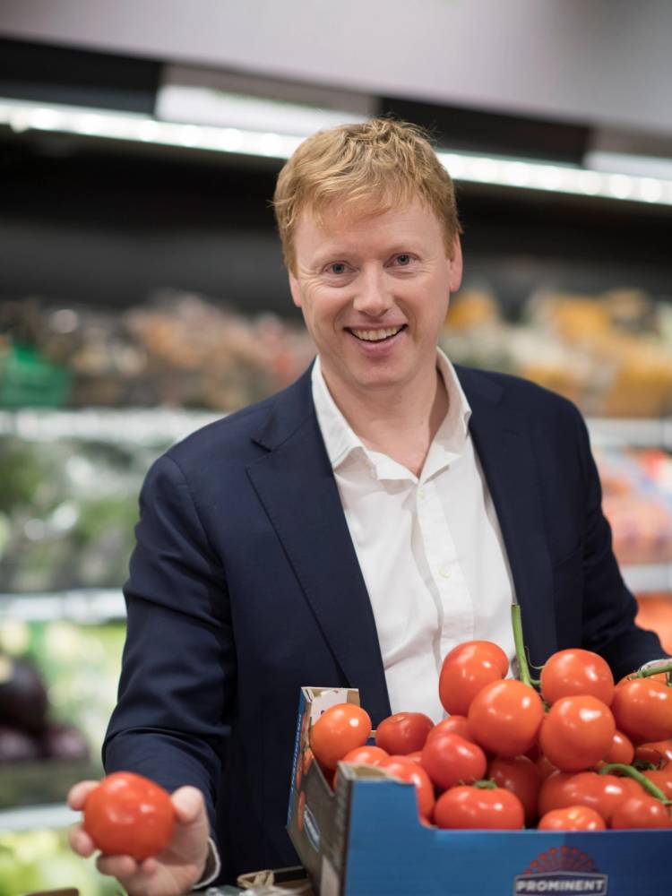 Person i dressjakke som handler tomater i en butikk, med grønnsaker i bakgrunnen.