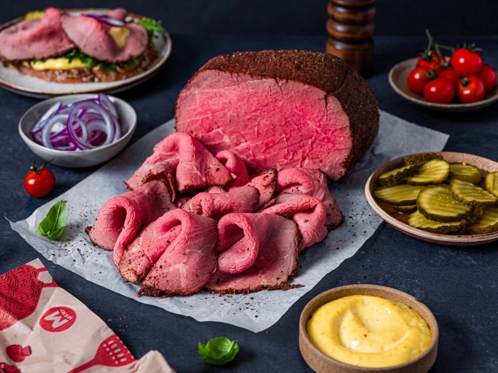 Skarpt skåret rosa og brunt kjøttstykke omgitt av små skåler med sylteagurk, løk og sennep, med en bunke med plakater i bakgrunnen.