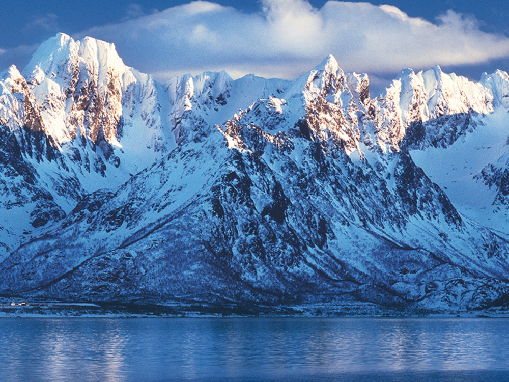 Snødekte fjell med speilblankt vann foran, under en delvis overskyet himmel.