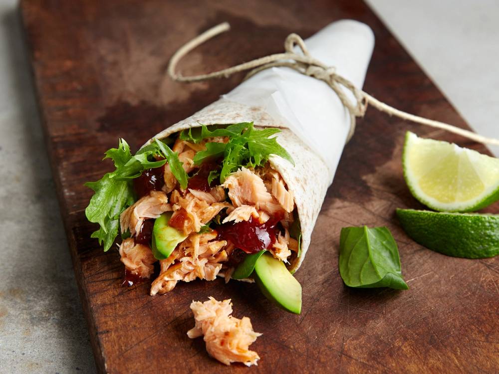 Pulled laks i en tortillawrap med grønnsaker og urter, med limebåter ved siden av på et treplate.