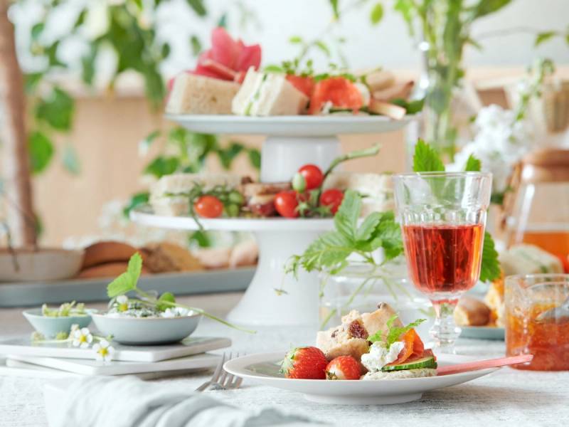 Et bord dekket med sommermat, inkludert kaker, frukt og en glass med rød drikke, med grønne planter i bakgrunnen.