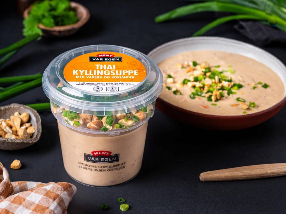 En kopp thai kyllingsuppe med vårløk og koriander ved siden av en pakke med samme suppe, på et svart bord med noen nøtter og en skje.