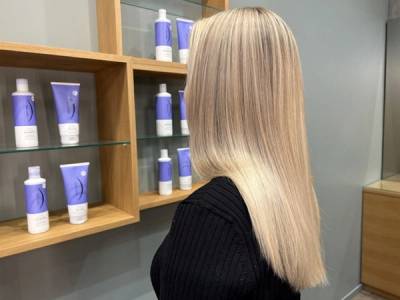 En person med langt, blondt hår står foran en vegg med hyller som har ulike hårprodukter, i en frisørsalong.