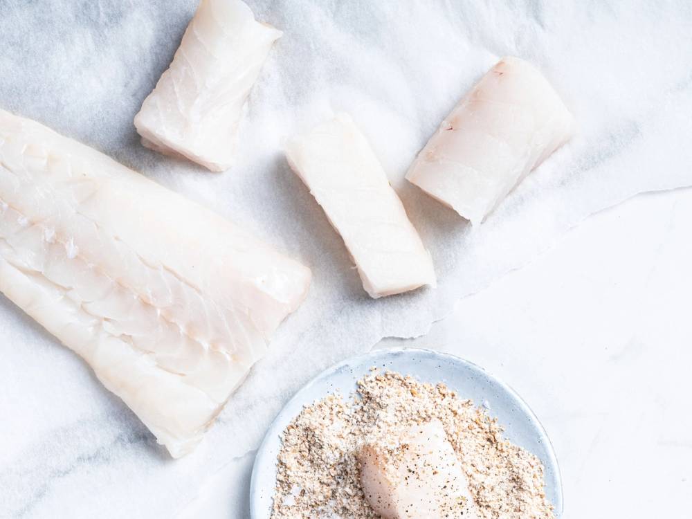 Fiskepinner og fiskefilet på et hvitt underlag med en skål med mel eller rasp, klar til matlaging.