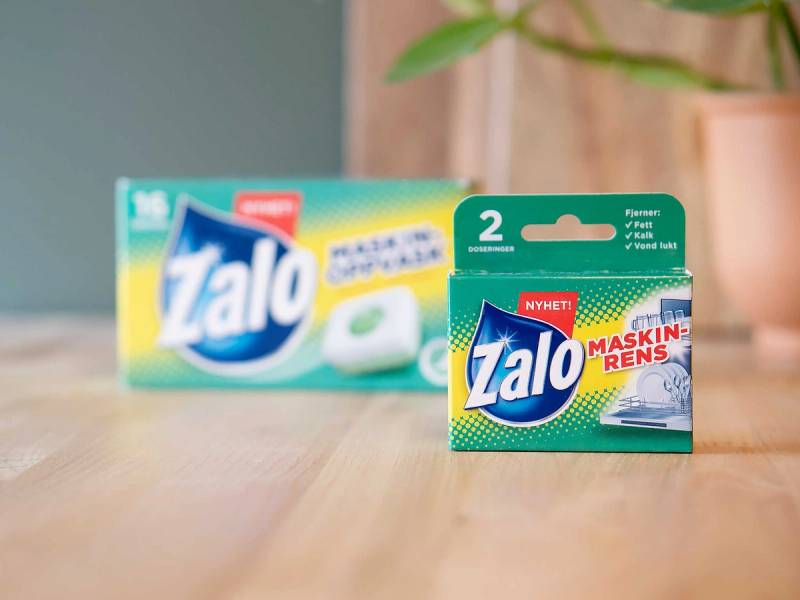 To pakker med Zalo-maskinrens, en større og en mindre, plassert på et bord med en plante i bakgrunnen.