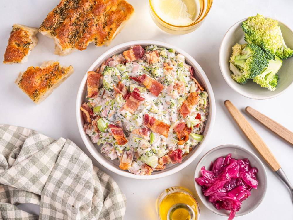 En salat med bacon, grønnsaker og ost i en stor bolle, omgitt av brokkoli, rødkål, brød og dressing på et hvitt bord