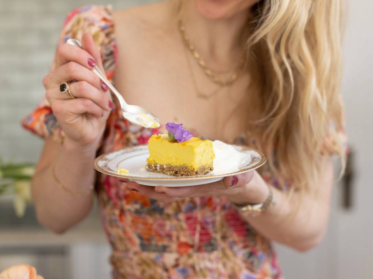 En person holder en tallerken med en skive av gul kake pyntet med lilla blomster og spiser med en skje, med en person i en blomstrete kjole i bakgrunnen.