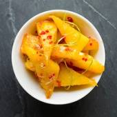 En skål med skivede, gule frukter som ser ut som ananas eller mango, pyntet med små røde krydderbiter og grønne urter.