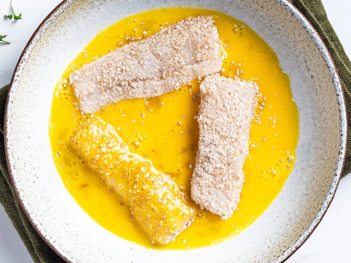 To fiskestykker dekkes i mel og ligger i en skål med vispet egg, klar for panering.